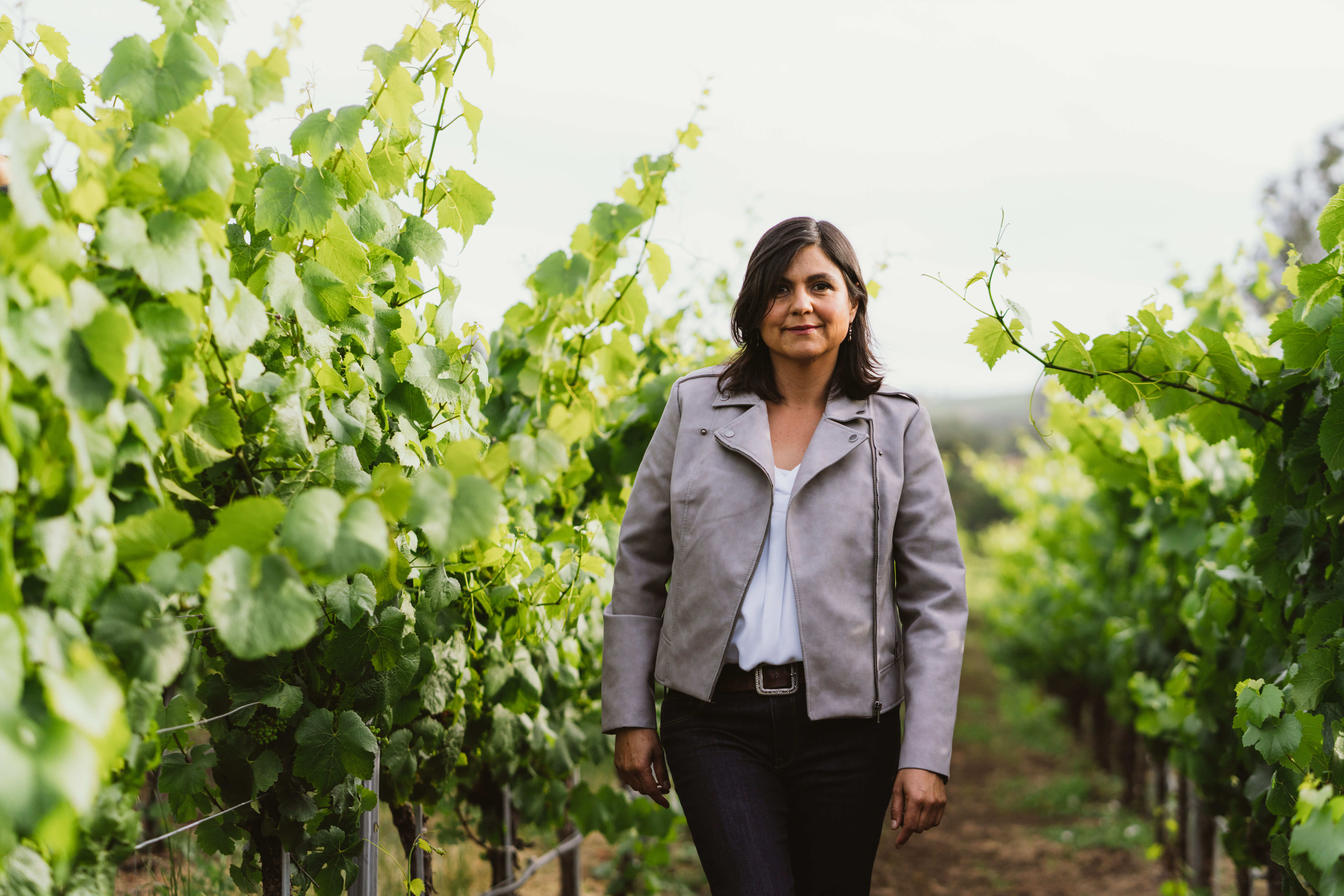 A photo of a woman with short brown hair in a grey jacket and black pants, standing in a lush green vineyard.